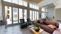 This thoughtfully designed single-floor space combines contemporary architecture with carefully curated interiors across several distinct areas. Soaring double-height ceilings define much of the layout, creating an expansive, airy atmosphere throughout. Large floor-to-ceiling glass doors and clerestory windows flood the interior with natural light while framing views of a lush outdoor courtyard complete with a fireplace and manicured landscaping.

The main lounge area features a sleek, wall-mounted linear fireplace set against light wood-panel surround, with a patterned encaustic tile runner anchoring the seating arrangement. Teal upholstered armchairs, a deep burgundy sectional, and a wood coffee table adorned with succulent arrangements compose an inviting and stylish gathering space.

A secondary lounge showcases a dramatic floor-to-ceiling wine display tower in dark metal framing, serving as both a functional feature and a striking architectural focal point. High-back upholstered booth seating with vibrant orange accent cushions, framed artwork, and warm wood-look tile flooring contribute to the refined yet comfortable ambiance.

The kitchen area is distinguished by bold, geometric encaustic tile covering both the floor and a full accent wall. Light wood cabinetry, grey stone countertops, a matte black faucet, and open geometric shelving complete this visually dynamic space.

A bright dining area with large windows overlooks the courtyard, offering views of tropical foliage and the surrounding community, enhanced by curated artwork and natural greenery centerpieces.