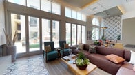 This thoughtfully designed single-floor space combines contemporary architecture with carefully curated interiors across several distinct areas. Soaring double-height ceilings define much of the layout, creating an expansive, airy atmosphere throughout. Large floor-to-ceiling glass doors and clerestory windows flood the interior with natural light while framing views of a lush outdoor courtyard complete with a fireplace and manicured landscaping.

The main lounge area features a sleek, wall-mounted linear fireplace set against light wood-panel surround, with a patterned encaustic tile runner anchoring the seating arrangement. Teal upholstered armchairs, a deep burgundy sectional, and a wood coffee table adorned with succulent arrangements compose an inviting and stylish gathering space.

A secondary lounge showcases a dramatic floor-to-ceiling wine display tower in dark metal framing, serving as both a functional feature and a striking architectural focal point. High-back upholstered booth seating with vibrant orange accent cushions, framed artwork, and warm wood-look tile flooring contribute to the refined yet comfortable ambiance.

The kitchen area is distinguished by bold, geometric encaustic tile covering both the floor and a full accent wall. Light wood cabinetry, grey stone countertops, a matte black faucet, and open geometric shelving complete this visually dynamic space.

A bright dining area with large windows overlooks the courtyard, offering views of tropical foliage and the surrounding community, enhanced by curated artwork and natural greenery centerpieces.