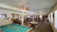 This single-story community space is thoughtfully designed to accommodate a variety of leisure and social activities. The open-plan layout is anchored by a full-size pool table with a green felt surface, making it a natural gathering point for recreation. Ceiling fans are mounted throughout, complementing the recessed lighting to maintain a comfortable and well-lit atmosphere across the entire floor.

The space is divided into distinct functional zones without the use of full walls, allowing for an airy, connected feel. Wicker and rattan seating arrangements are grouped around glass-topped tables, creating intimate areas ideal for conversation or casual gatherings. Toward the rear of the space, a library area is visible, lined with bookshelves and accompanied by computer workstations, offering a quieter retreat for reading or research.

Large windows fitted with slatted blinds line the walls, allowing natural light to filter in while maintaining privacy. Support columns are integrated into the layout, subtly defining different areas without interrupting the open flow. The neutral color palette of the walls and flooring provides a calm, welcoming backdrop that ties all the zones together, making this a versatile and inviting communal environment suitable for a wide range of activities and group sizes.