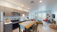 This thoughtfully designed single-floor residence offers a seamless blend of modern aesthetics and functional living. The open-concept layout connects the kitchen and living area, creating a bright and inviting atmosphere. The kitchen is equipped with stainless steel appliances, including a refrigerator, electric range, microwave, and dishwasher, complemented by two-tone cabinetry in warm wood tones and dark base cabinets. Light quartz countertops and recessed lighting add a clean, contemporary finish, while a butcher-block island with bar seating provides a casual dining option. Light wood-look flooring runs throughout the main living areas, enhancing the sense of openness. The living room benefits from abundant natural light streaming through large sliding glass doors that open to an outdoor view, and features ample space for comfortable seating arrangements. The bedroom is carpeted for warmth and comfort, offering a generous layout with a large window dressed in elegant draping curtains that allow soft, natural light. The bathroom maintains the modern design language with a floating wood-tone vanity, oval mirror, and a shower/tub combination. Throughout the space, clean white walls and recessed lighting contribute to a bright, airy ambiance ideal for contemporary living.