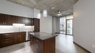 This thoughtfully designed one-bedroom, one-bathroom unit showcases a contemporary industrial aesthetic throughout its well-proportioned single-floor layout. The defining architectural feature is the exposed concrete ceiling, which carries through the open-plan living and kitchen areas, lending an urban loft character to the space.

The kitchen is a standout element, outfitted with rich dark walnut cabinetry, a textured tile backsplash, and a full suite of stainless steel appliances including a refrigerator, range, microwave, and dishwasher. A central island with a gray quartz-style countertop provides additional workspace and casual seating. Decorative pendant lights hang above the island, complementing the ceiling fan that anchors the adjacent living area. Floor-to-ceiling sliding glass doors lead to a private balcony, filling the main living space with natural light and outdoor views.

The bedroom is carpeted for comfort, features its own ceiling fan, an exposed concrete ceiling, and a large window. Two interior doors lead to a dedicated walk-in closet with wrap-around hanging rods and a full bathroom. The bathroom is finished with a glass-enclosed walk-in shower clad in large white subway tiles, a dark-framed vanity mirror, and a single undermount sink with a quartz countertop, maintaining the unit's cohesive modern palette throughout.