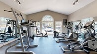 Located in Riverdale, GA, this dedicated fitness center offers a well-equipped workout space designed to support a range of exercise routines. The room features a striking vaulted ceiling that lends an open, airy feel despite the space's modest footprint, while track lighting above ensures the area is well-illuminated throughout.

A prominent arched window flanked by rectangular windows along the front wall allows abundant natural light to flood the interior, offering views of the surrounding wooded landscape and providing a refreshing atmosphere during workouts. A full-length wall mirror on one side visually expands the space and assists users in monitoring their form.

The fitness center is outfitted with a comprehensive selection of equipment, including treadmills, elliptical trainers, a stationary recumbent bike, a rowing machine, and a multi-function cable/weight machine for strength training. Durable rubber flooring covers the entire space, providing safety and impact absorption. A French door entry with flanking windows adds a refined architectural touch to the rear wall, which is accented by neutral warm-gray tones throughout. A decorative potted plant near the entrance adds a subtle natural element to the otherwise functional environment. Fitness center rules are posted on the wall for user reference.