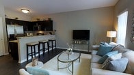 This two-storey space offers a thoughtfully designed interior that blends contemporary style with functional living. The main floor showcases an open-concept layout where the kitchen flows seamlessly into the living area, creating a connected and sociable environment. The kitchen is fitted with dark espresso cabinetry, stainless steel appliances, and a granite-topped breakfast bar with seating for several stools — making it ideal for casual dining and entertaining. The living area is anchored by dark hardwood flooring and soft neutral grey walls, providing a versatile backdrop for the space. A plush white area rug defines the seating zone, complemented by a glass-topped oval coffee table with a metallic frame. The room is accessorized with teal accent cushions, decorative vases, and subtle greenery atop the kitchen cabinetry, adding warmth and character. A flat-screen television is mounted on a sleek media stand positioned against the main wall. The overall ambiance is modern and polished, with a cohesive colour palette of grey, white, and dark tones throughout. The second floor likely accommodates the private living quarters of the home.