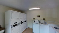 This single-floor shared laundry facility is a practical and well-organized space designed to accommodate the everyday laundry needs of its users. The room features a clean, utilitarian layout with white-painted walls and a neutral tile floor that is easy to maintain. Overhead fluorescent lighting ensures the space is well-lit throughout.

Along one wall, a bank of stacked commercial-style dryers is arranged in a neat row, maximizing capacity within the available footprint. Opposite, a row of top-loading washing machines lines the adjacent wall, providing ample washing capacity. The machines are standard commercial units, each equipped with coin or card-operated controls, making them accessible and straightforward to use. Informational and instructional signage is posted on the walls to guide users on proper machine operation and facility etiquette.

The overall ambiance of the space is clean and no-frills, prioritizing function and efficiency. The white cabinetry and appliances contribute to a bright, orderly atmosphere. With its compact yet well-planned arrangement, this laundry room delivers a convenient, communal laundering solution within a tidy and well-maintained environment.