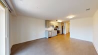 This single-floor unit is designed with a clean, contemporary aesthetic and an efficient open-concept layout. The living area is generously proportioned, featuring wood-look flooring in a neutral tone that extends throughout the space, crisp white walls, and flush-mount ceiling lighting. A sliding glass door opens to a balcony, providing outdoor access and filling the room with natural light and views of surrounding greenery.

The kitchen is positioned along one wall and integrates seamlessly with the living space. It is outfitted with gray-toned shaker-style cabinetry, light granite-look countertops, and a full suite of stainless steel appliances including a refrigerator, gas range, microwave, and dishwasher. A single-basin sink is set beneath upper cabinetry, maximizing storage and functionality.

A hallway leads from the main living area toward the bathroom and additional rooms. The bathroom features a soaking tub and shower combination with classic white subway tile accented by a decorative horizontal tile inset. Light wood-look tile flooring, a white vanity with a solid countertop, and a large mirror complete the clean, bright finish. Throughout the unit, the design prioritizes a neutral, move-in-ready atmosphere.