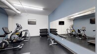 This single-level fitness room is designed to provide a practical and focused workout environment. The space features a clean, modern aesthetic with white walls accented by a cool blue feature wall that adds a sense of depth and visual interest. Durable dark rubber flooring covers the entire area, offering both safety and functionality for a range of exercises.

A full dumbbell rack lines the center of the room, offering a comprehensive range of weights to accommodate various strength-training routines. A flat workout bench sits nearby, providing a versatile surface for multiple exercises. Cardio equipment, including an elliptical machine, is positioned along the perimeter, maximizing the usable floor space.

A large floor-to-ceiling mirror spans the length of the blue accent wall, effectively doubling the perceived size of the room while allowing users to monitor their form during workouts. Flat-screen televisions are mounted on both the mirrored and opposing walls, offering entertainment options during sessions. Overhead fluorescent lighting ensures the space is bright and well-illuminated throughout.