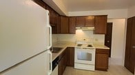 This two-story residence includes a well-equipped kitchen that serves as a central feature of the home. The kitchen is outfitted with rich, dark wood cabinetry that extends to the upper walls, providing generous storage space. White appliances, including a full-size refrigerator, an electric range with coil burners, and a built-in dishwasher, offer everyday functionality. A ventilation hood is mounted above the range, and light-toned countertops contrast neatly with the darker cabinet finish. The tile flooring adds durability and easy maintenance to the space. The layout follows an efficient L-shaped configuration, optimizing workflow between the cooking, prep, and storage zones. Under-cabinet lighting adds warmth to the kitchen area. The two-floor layout of the residence suggests additional living, sleeping, or utility spaces distributed across both levels, offering separation between common and private areas. Overall, the space presents a practical and organized interior with a classic design sensibility rooted in functionality.