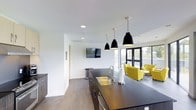 This single-floor space showcases a sleek, modern design that seamlessly blends a functional kitchen area with an inviting lounge. The open-concept layout promotes a sense of spaciousness and connectivity throughout the interior.

The kitchen island takes center stage, featuring a dark countertop, an undermount double sink with a contemporary faucet, and dark cabinetry with stainless steel hardware. Three striking black pendant lights hang above the island, adding both style and focused illumination. Recessed ceiling lighting further brightens the crisp white ceilings and walls, contributing to the clean, minimalist atmosphere.

Adjacent to the kitchen, the lounge area is furnished with bold yellow-green accent chairs that inject a lively pop of color against the neutral backdrop. A wall-mounted flat-screen television is positioned for comfortable viewing from multiple areas of the space.

Large floor-to-ceiling windows and glass doors along the far wall allow abundant natural light to flood the interior while offering views of the outdoor surroundings. The flooring throughout features a warm wood-tone finish that grounds the space and complements the contemporary design elements. Together, these features create a well-appointed, multi-functional environment ideal for both gathering and everyday use.