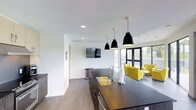 This single-floor space showcases a sleek, modern design that seamlessly blends a functional kitchen area with an inviting lounge. The open-concept layout promotes a sense of spaciousness and connectivity throughout the interior.

The kitchen island takes center stage, featuring a dark countertop, an undermount double sink with a contemporary faucet, and dark cabinetry with stainless steel hardware. Three striking black pendant lights hang above the island, adding both style and focused illumination. Recessed ceiling lighting further brightens the crisp white ceilings and walls, contributing to the clean, minimalist atmosphere.

Adjacent to the kitchen, the lounge area is furnished with bold yellow-green accent chairs that inject a lively pop of color against the neutral backdrop. A wall-mounted flat-screen television is positioned for comfortable viewing from multiple areas of the space.

Large floor-to-ceiling windows and glass doors along the far wall allow abundant natural light to flood the interior while offering views of the outdoor surroundings. The flooring throughout features a warm wood-tone finish that grounds the space and complements the contemporary design elements. Together, these features create a well-appointed, multi-functional environment ideal for both gathering and everyday use.