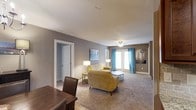 This single-floor space offers a well-appointed and inviting interior designed for comfortable everyday living. The living area is a standout feature, showcasing plush wall-to-wall carpeting that adds warmth underfoot and a soft, welcoming ambiance throughout. Neutral gray walls provide a versatile backdrop, complemented by crisp white trim and ceilings that brighten the space. A ceiling fan overhead offers both practical airflow and a touch of casual elegance. Natural light filters generously into the room through a set of glass doors dressed with vibrant teal curtains, adding a pop of color and personality to the otherwise neutral palette. The living area is thoughtfully arranged with a mustard yellow sofa and armchair combination, a round coffee table, and coordinating accent lamps that create a warm, layered lighting scheme. Adjacent to the living space, a dining area with a dark wood table and chairs provides a defined space for meals and gatherings. A doorway on the far side of the room leads to additional areas of the home, hinting at a functional and connected floor plan. The overall atmosphere is cozy, modern, and move-in ready.