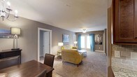 This single-floor space offers a well-appointed and inviting interior designed for comfortable everyday living. The living area is a standout feature, showcasing plush wall-to-wall carpeting that adds warmth underfoot and a soft, welcoming ambiance throughout. Neutral gray walls provide a versatile backdrop, complemented by crisp white trim and ceilings that brighten the space. A ceiling fan overhead offers both practical airflow and a touch of casual elegance. Natural light filters generously into the room through a set of glass doors dressed with vibrant teal curtains, adding a pop of color and personality to the otherwise neutral palette. The living area is thoughtfully arranged with a mustard yellow sofa and armchair combination, a round coffee table, and coordinating accent lamps that create a warm, layered lighting scheme. Adjacent to the living space, a dining area with a dark wood table and chairs provides a defined space for meals and gatherings. A doorway on the far side of the room leads to additional areas of the home, hinting at a functional and connected floor plan. The overall atmosphere is cozy, modern, and move-in ready.