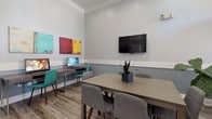 This single-floor space functions as a versatile meeting and business center room, thoughtfully designed to accommodate both collaborative gatherings and individual work. The room features elevated ceilings adorned with crown molding, lending a sense of height and architectural refinement. Walls are finished in a two-tone scheme, with a crisp white upper portion and a cool grey lower half divided by a clean chair rail, creating a polished and professional atmosphere.

The flooring consists of light wood-tone planks that run throughout, adding warmth to the otherwise neutral palette. Recessed lighting and track lighting provide even illumination across the room. A wall-mounted flat-screen television serves as a central focal point, ideal for presentations or collaborative viewing.

A rectangular conference table surrounded by upholstered grey chairs occupies the center of the room, accommodating multiple participants comfortably. Along one wall, a dedicated workstation desk is equipped with a desktop computer and teal accent chairs, offering a practical workspace for individual use. Colorful abstract artwork and potted greenery add visual interest and a welcoming touch to the overall environment.