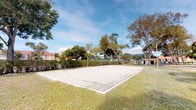 This outdoor recreational area centers around a well-maintained sand volleyball court, clearly delineated by white boundary lines and equipped with a regulation-height net strung between sturdy metal poles. The sandy playing surface is surrounded by a generous lawn of green grass, providing ample space around the court for spectators or additional activities. Mature trees frame the perimeter of the space, offering natural shade and a sense of enclosure while maintaining an open, airy feel beneath the expansive blue sky. A neatly trimmed hedge line runs along one side of the area, providing a natural boundary and added privacy. Residential structures are visible in the background, suggesting this court is part of a community recreational amenity. The overall setting is bright and inviting, benefiting from abundant natural light throughout the day. The combination of the sand court, surrounding greenery, and tree canopy creates a casual yet functional outdoor environment well-suited for recreational play and social gatherings.