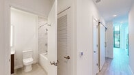 This thoughtfully designed single-floor unit offers a contemporary living environment with a clean, minimalist aesthetic throughout. The open layout maximizes the available footprint, with light wood flooring running continuously through the main living and kitchen areas to create a cohesive, airy feel.

The kitchen is outfitted with flat-front cabinetry in a muted blue-gray finish, white countertops, stainless steel appliances including a built-in microwave, electric range, dishwasher, and refrigerator, and a single-basin sink with a brushed gold faucet. Wall-mounted open shelving adds additional storage and visual interest above the counter line.

The living area benefits from dramatic floor-to-ceiling windows framed in dark metal, allowing abundant natural light and offering elevated views of the surrounding urban streetscape and neighboring rooftops. A secondary room features its own window access and includes a built-in closet with sliding doors, dual hanging rods, and integrated shelving for organized storage.

The bathroom is finished with large-format marble-look wall tiles in the tub-shower surround, complemented by a floating vanity with a backlit mirror, white countertop, and neutral floor tiles. A sliding barn-style door accents the bathroom entry, adding a modern architectural detail to the overall design.