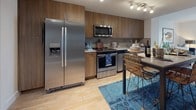 This single-floor residence offers a thoughtfully designed layout that balances modern aesthetics with functional living. Light hardwood flooring flows throughout, unifying the open living spaces with a warm, cohesive feel.

The living room is a comfortable retreat, centered around a plush navy blue sofa paired with a geometric-patterned area rug. Framed abstract artwork anchors the space above the seating area, while a mix of floor and table lamps provides layered ambient lighting. The room's neutral white walls keep the space feeling bright and airy.

The kitchen is outfitted with warm wood cabinetry, a textured mosaic tile backsplash, and a full suite of stainless steel appliances including a side-by-side refrigerator, range, microwave, and dishwasher. A high-top dining table with woven rattan chairs sits alongside the kitchen, illuminated by a sleek track lighting system overhead, making the area ideal for both casual dining and entertaining.

The bedroom features a bold teal accent wall that adds depth and character to the room. A large window with access to a balcony invites natural light and outdoor views, while plush carpeting underfoot adds comfort. The adjacent bathroom is finished with wood-toned vanity cabinetry, a clean white sink, and light hardwood-style flooring, maintaining the residence's consistent modern design language throughout.