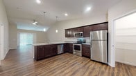 This single-floor residence at Redwood Norwalk in Norwalk, Ohio, offers a thoughtfully designed layout with modern finishes throughout. The open-concept kitchen is a standout feature, showcasing rich dark cabinetry, granite-look countertops, stainless steel appliances including a refrigerator, electric range, and built-in microwave, and durable wood-look vinyl plank flooring. Pendant lighting accents the kitchen island/peninsula, which flows seamlessly into the living area. The living room benefits from vaulted ceilings and natural light from large windows, creating an airy, open ambiance, with wood-look flooring continuing underfoot. Bedrooms are carpeted for comfort and are bright with natural light from multiple windows, each equipped with a ceiling fan. One bedroom provides direct sightlines to the kitchen's pendant lighting, reinforcing the open feel of the floor plan. A walk-in pantry or closet with shelving is accessible from the kitchen area. The bathrooms feature wood-look vinyl flooring, dark vanity cabinetry with granite-look countertops, and clean white fixtures — one with a tub/shower combo and another with a walk-in shower. The overall design presents a cohesive, contemporary aesthetic with neutral tones throughout.