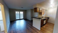 This single-story space presents an open-concept design that seamlessly connects the kitchen and living areas. The living space is finished with rich, dark hardwood flooring that extends throughout, creating a cohesive and grounded aesthetic. A set of rear-facing double doors with shuttered windows allows natural light to filter into the living area, enhancing the sense of openness.

The kitchen is positioned to one side and features warm-toned wood cabinetry with upper and lower storage, dark granite-style countertops, and a peninsula that provides a natural division between the kitchen and living space while offering additional counter workspace. A sink is integrated into the peninsula, adding functional convenience. The appliances are finished in white and black tones, complementing the cabinetry. A window above the counter area brings additional natural light into the kitchen.

The neutral wall tones throughout the space contribute to a clean, versatile backdrop, while the open floor plan allows for flexible furniture arrangement. The overall ambiance is practical and functional, suited for everyday living.