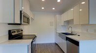 This single-floor space showcases a thoughtfully designed, modern interior with a strong emphasis on clean lines and a minimalist aesthetic. The kitchen is a standout feature, outfitted with high-gloss white cabinetry that extends from floor to ceiling, maximizing storage while maintaining a streamlined, uncluttered look. Stainless steel appliances, including a built-in microwave, an electric range, and a dishwasher, lend a professional and polished feel to the space. The kitchen also features light-toned quartz countertops that complement the white cabinetry beautifully, along with a deep undermount sink fitted with a contemporary brushed nickel faucet. Recessed lighting is installed throughout the ceiling, providing consistent, bright illumination across the entire kitchen area. The flooring consists of durable wood-look planks in a warm medium-brown tone, adding a subtle sense of warmth that contrasts elegantly with the predominantly white palette. The walls are finished in a crisp, clean white, contributing to the overall bright and airy ambiance. The layout is highly functional, with a galley-style configuration that optimizes workflow and efficiency in the kitchen space.