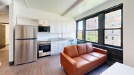 Located at 618 Clyde St in Pittsburgh's Oakland neighborhood, this two-bedroom, two-bathroom unit offers a functional and modern layout across a single floor. The open-concept kitchen and living area serves as the heart of the space, featuring white shaker-style cabinetry, gray countertops, stainless steel appliances including a refrigerator, range, dishwasher, and microwave, along with wood-style flooring. Large, dark-framed windows flood the area with natural light and provide views of the surrounding Pittsburgh streetscape and greenery. The two bedrooms are carpeted and well-sized, each equipped with built-in furnishings including a bed frame with integrated nightstands. The primary bedroom features distinctive dark wainscoting panels and multiple windows, while the second bedroom includes a dedicated study desk area with city views. The unit includes two full bathrooms: one with a tiled tub-shower combo, white vanity, and large-format tile flooring, and a second with a walk-in shower stall and matching tile flooring. Two hallways connect the rooms efficiently throughout the unit. The overall design is clean and contemporary, making it a practical and comfortable residence in the heart of Pittsburgh.