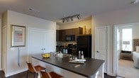 This single-floor residence offers a thoughtfully designed open-concept layout that maximizes both functionality and style. The kitchen serves as the focal point of the space, showcasing rich dark wood cabinetry that extends to the upper walls, providing ample storage while lending a warm, contemporary aesthetic. Black appliances, including a refrigerator, range, and built-in microwave, complement the cabinetry seamlessly for a cohesive, modern look. A generously sized kitchen island anchors the space, topped with a dark stone-style countertop that offers plenty of prep space as well as casual bar seating with leather-accented chairs — ideal for dining or entertaining. The island also features a stainless steel sink, adding convenience to the layout. Overhead, track lighting illuminates the kitchen with warm, directional light, highlighting the space's design details. The surrounding walls are finished in a clean, neutral tone, enhancing the brightness of the interior. Adjacent doors hint at well-connected living areas and closet storage, contributing to the overall practical flow of the residence. The space balances modern design elements with everyday livability.