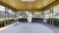 This outdoor courtyard gazebo serves as a welcoming communal gathering space, thoughtfully designed for relaxation and social interaction. The structure features a solid overhead canopy with a warm golden-toned ceiling, providing ample shade while maintaining an open, airy feel. Decorative wrought iron lattice panels with intricate floral and vine motifs frame the perimeter of the gazebo, adding an elegant architectural character to the space.

The concrete flooring is spacious enough to accommodate multiple seating arrangements, and the area is currently furnished with black metal benches, chairs, and a small side table — offering comfortable spots for residents to gather and unwind. A low ornamental railing borders the gazebo, defining the space while keeping sightlines open to the surrounding landscape.

From the gazebo, visitors enjoy unobstructed views of a well-maintained courtyard with neatly trimmed hedgerows, mature trees, and a concrete pathway that leads through lush green lawns toward the surrounding multi-story brick buildings. The natural greenery and tree canopy visible from the seating areas contribute to a calm, park-like ambiance. The overall setting balances functional outdoor living with classic decorative detailing, making this courtyard a pleasant and inviting communal retreat.