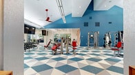 Located at 1500 Chase Arbor Common in Virginia Beach, VA, this fitness center is a dynamic, single-floor exercise facility designed to support a full spectrum of workout routines. The space makes a bold visual statement with its striking geometric-patterned flooring in blue, white, and gray triangles, complemented by vivid blue accent walls and bold red-and-white diagonal stripe details that energize the environment throughout.

The main workout area is generously proportioned, featuring soaring vaulted ceilings in key sections that contribute to an open, airy atmosphere. A row of floor-to-ceiling glass walls and large windows along one side floods the interior with abundant natural light while offering views of the surrounding greenery. Red ceiling fans add both a functional and stylistic accent overhead.

The facility is comprehensively equipped with a full suite of strength machines, free weights, adjustable benches, and cable systems. A dedicated cardio zone houses stationary bikes and additional aerobic equipment. A standout feature is the Treadwall climbing machine, situated in a bright, windowed corner alcove that opens toward the outdoors, providing a unique and engaging fitness option. Crash mats and open floor space further support functional training and stretching. Mounted flat-screen televisions and recessed lighting complete the modern, well-appointed interior.