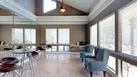 This single-floor space makes a strong architectural impression with its dramatic vaulted ceiling, which rises to a striking peak and is complemented by a raised tray ceiling detail below, adding depth and visual interest overhead. A central pendant light fixture hangs from the apex, providing both functional illumination and a decorative focal point.

Natural light is a defining characteristic of this space. Multiple large windows fitted with horizontal blinds line the walls, while a skylight positioned near the roofline further enhances the bright, airy atmosphere. The windows offer views of the surrounding greenery outside, connecting the interior to the outdoors.

The flooring consists of neutral-toned large-format tiles that extend throughout, contributing to the clean, open feel of the room. The warm taupe wall color pairs well with the white crown molding and window trim, creating a cohesive and polished aesthetic.

The space is furnished with a mix of seating options, including a tall bistro-style table with modern bar stools positioned near a mirrored wall, and a pair of houndstooth-patterned accent chairs arranged in a conversational layout. A built-in ledge along one wall provides surface space, currently housing a printer, suggesting utility alongside comfort.