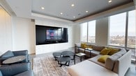 This sophisticated media room is designed with both comfort and functionality in mind. At the heart of the space is a striking floor-to-ceiling built-in entertainment wall finished in crisp white cabinetry, framing a large flat-screen television and providing ample concealed storage. The tray ceiling adds architectural depth and dimension to the room, while recessed lighting ensures a well-lit, versatile atmosphere suited for both viewing and socializing. Expansive floor-to-ceiling windows flood the room with natural light and offer sweeping views of the surrounding cityscape, creating a bright and airy ambiance that complements the clean, contemporary aesthetic. Seating is arranged thoughtfully around a set of sleek, clustered circular coffee tables atop a textured area rug, anchoring the space with a warm, layered feel. A generous sectional sofa in neutral tones is accented with bold mustard-yellow throw pillows, introducing a vibrant pop of color against the otherwise muted palette of grays and whites. The overall design strikes a refined balance between a high-performance entertainment environment and an inviting, stylish lounge space.