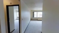 This single-floor space presents a straightforward, functional layout with a clean and neutral aesthetic throughout. The main living area is carpeted in light grey, providing a soft, understated foundation that complements the white walls. A textured ceiling adds subtle dimension to the space overhead.

A large window in the living area frames elevated city views, allowing natural light to fill the room and offering a glimpse of surrounding urban architecture. A baseboard heating unit is positioned beneath the window, providing practical climate control. The space is accessed through a corridor that connects to an adjoining room, also featuring its own window for additional natural light.

The interior walls are painted in a crisp white with contrasting dark-trimmed doorframes, lending a clean, defined structure to the layout. The overall condition of the space appears well-maintained and move-in ready, offering a blank canvas for a variety of uses. The open, uncluttered design allows for flexible furniture arrangements, making it adaptable to different lifestyle needs. The elevated position of the unit, as suggested by the city views, adds an appealing sense of height and openness to the overall ambiance.