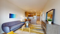 This single-floor unit showcases a cohesive modern design anchored by light hardwood flooring that flows seamlessly through every room. The open-concept living area serves as the heart of the space, featuring a mid-century-inspired seating arrangement with a slate-blue sofa, a bold yellow accent chair, and a striped area rug in gold and grey tones. Colorful wall art adds a vibrant, curated touch to the neutral palette.

The kitchen is thoughtfully designed with warm wood cabinetry extending from floor to ceiling, stainless steel appliances including a range, microwave, dishwasher, and refrigerator, and a clean white countertop. A peninsula with bar seating connects the kitchen to the living area, making it ideal for both casual dining and entertaining. Pendant lighting over the peninsula adds a refined finishing detail.

The bedroom is a calm retreat, benefiting from a large floor-to-ceiling window that draws in natural light and frames outdoor views. A ceiling fan enhances comfort, while art on the wall and lush greenery contribute warmth.

The bathroom is finished with a wooden vanity, open wood shelving, a wide mirror, and a sliding glass walk-in shower, maintaining the space's consistent warm, contemporary aesthetic throughout.