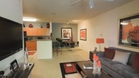 This single-floor residence showcases an open-concept layout that connects the kitchen, dining, and living areas into one cohesive, flowing space. The kitchen features warm wood cabinetry, a dark countertop, and a white breakfast bar that acts as a natural divider between the cooking and dining zones. Track lighting runs along the ceiling, supplementing the recessed lights to ensure the space is well-illuminated throughout. The dining area accommodates a sleek table with black bar-style chairs, positioned beneath additional track lighting. Bold red and black framed artwork adorns the accent wall, adding visual interest and tying together the space's color palette of red, black, and gray. The living area is anchored by a gray sectional sofa adorned with deep red and charcoal accent pillows, complemented by a dark coffee table with decorative candles. Light-toned carpeting covers the floors in the main living areas, contributing to a warm, comfortable ambiance. The overall aesthetic is modern and cohesive, with consistent use of bold accent colors and contemporary furnishings creating a stylish and functional environment.