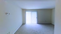 This single-floor space features a clean and straightforward layout designed for comfortable everyday living. The main living area is finished with wall-to-wall beige carpeting, providing a neutral and cohesive foundation throughout. Smooth, white-painted walls and a flat ceiling contribute to the room's bright, open feel.

A prominent sliding glass door serves as a focal point of the living area, fitted with full-length vertical blinds that offer privacy control while still allowing generous natural light to permeate the space. The door provides direct access to an exterior area, enhancing the sense of openness and connection to the outdoors.

White baseboards run along the perimeter of the room, adding a clean, finished detail to the overall aesthetic. Electrical outlets are positioned at convenient intervals along the walls. The space presents a blank canvas, well-suited for a variety of furniture arrangements and personal touches. Overall, the space offers a functional and well-lit environment with a simple, understated design.