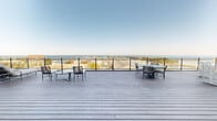 Located at 233 Lake Ave in Racine, Wisconsin, this rooftop terrace is a standout outdoor destination defined by its generous open-air deck space and breathtaking panoramic views of Lake Michigan and the neighboring marina. The deck is finished with wide-plank composite decking in a cool gray tone, lending a clean, modern aesthetic throughout. A sleek glass-panel railing system runs along the perimeter, preserving unobstructed sightlines to the water and horizon beyond. Multiple seating arrangements are distributed across the terrace, including dining sets, lounge chairs, and casual conversation groupings, accommodating a variety of uses from relaxed sunbathing to outdoor entertaining. A glass-fronted interior structure anchors one end of the terrace, featuring floor-to-ceiling glazing and glass doors that connect indoor and outdoor spaces seamlessly. The building's exterior cladding features contemporary horizontal siding that complements the deck's neutral palette. Views from the terrace extend across the marina's boat slips, the Racine lakefront, and out to the open waters of Lake Michigan, making for a dramatic backdrop at any time of day. The space is well-suited for both quiet reflection and social gatherings, offering an elevated outdoor experience above the Lake Avenue corridor.