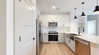 This thoughtfully designed one-bedroom, one-bathroom unit offers a clean, contemporary aesthetic throughout its single-floor layout. The open-concept kitchen and living area serves as the heart of the home, featuring light wood-look flooring, crisp white shaker-style cabinetry with matte black hardware, and stainless steel appliances including a French door refrigerator, electric range, and over-the-range microwave. A large peninsula with a white quartz countertop and undermount sink provides both workspace and casual dining potential, illuminated by a trio of sleek black pendant lights. A herringbone-pattern tile backsplash adds a subtle decorative touch. The living area benefits from natural light through a window with horizontal blinds. The bedroom offers a cozy atmosphere with soft carpeting, a ceiling fan, and a generously sized window. Two doors lead directly to a dedicated closet with built-in shelving and a hanging rod, and to the full bathroom. The bathroom is elegantly finished with wood-look flooring, a grey vanity with white countertop, a framed rectangular mirror, matte black fixtures, and a deep soaking tub with marble-style tile surround. A hallway connects the spaces with built-in open shelving, adding convenient storage throughout this well-appointed unit.
