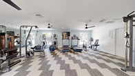 Located at 1119 Craftsman Dr in Virginia Beach, VA, this dedicated fitness center offers a well-appointed, single-floor workout space designed to support a wide range of exercise routines. The interior is finished with soft blue-gray walls and crisp white ceilings accented by crown molding, creating a clean and inviting atmosphere. A bold chevron-patterned carpet tile floor adds a modern visual element throughout the space.

The fitness center is generously equipped with multiple cardio machines, including treadmills, ellipticals, and a stationary bike, all positioned to take advantage of the natural light streaming in through several windows that offer views of the surrounding outdoor greenery. A wall-mounted flat-screen television is positioned for use during cardio workouts.

Strength training options include a multi-function cable machine, a weight bench, and a medicine ball rack, providing a comprehensive range of resistance exercise options. Full-length mirrors along one wall visually expand the space and allow users to monitor their form. A cable crossover/functional trainer unit is also available for versatile strength training.

Multiple ceiling fans ensure comfortable air circulation throughout the room. Additional amenities include a water fountain station and posted facility guidelines. The space is well-lit with a combination of natural and recessed lighting, making it a practical and pleasant environment for daily fitness activities.