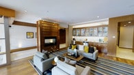 This well-appointed single-floor space showcases a seamless blend of modern design and warm, natural materials. At the heart of the living area stands a dramatic floor-to-ceiling wood-paneled fireplace, serving as a bold architectural focal point that anchors the room with both style and warmth. The open layout connects the living space to a sleek wet bar or kitchenette area, which features rich wood cabinetry with glass-front uppers, stone countertops, and integrated appliances — ideal for entertaining.

The living area is thoughtfully furnished with a generous arrangement of contemporary sofas and lounge chairs in neutral grey tones, accented by vibrant yellow and charcoal throw pillows that add a lively pop of color. A striped area rug in blue, grey, and cream tones defines the seating zone and complements the light hardwood flooring that runs throughout the space.

Recessed lighting illuminates the interior with a warm, even glow, while clean white ceilings and subtle architectural details contribute to a polished, upscale ambiance. The overall atmosphere is sophisticated yet inviting, combining functional design with curated aesthetic choices that make this space ideal for both relaxation and social gatherings.