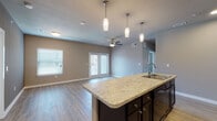 This single-floor residence offers a thoughtfully designed open-concept layout that seamlessly connects the kitchen, dining, and living areas. The kitchen serves as the centerpiece of the space, anchored by a large island with a granite-style countertop and an integrated sink, complemented by sleek dark cabinetry, black appliances including a refrigerator, electric range, microwave, and dishwasher, and a tasteful tile backsplash. Pendant lighting over the island adds a refined touch, while recessed ceiling lights provide ample illumination throughout.

The open living area features warm hardwood-style flooring, neutral-toned walls, and a ceiling fan, creating a comfortable and airy atmosphere. French doors lead to a private balcony or patio, allowing natural light to pour in. A secondary bedroom is finished with plush carpet, dual windows with blinds, and its own ceiling fan, offering a quiet retreat.

The bathroom is well-equipped with a dark-cabineted vanity topped with a granite-style countertop, a full tub-shower combination with tiled surround, and hardwood-style flooring. Throughout the space, consistent design choices — including neutral grey-toned walls, white trim, and coordinated finishes — contribute to a clean, modern, and cohesive aesthetic.