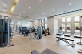 Located at 3784 West 88th Street in Hialeah, FL, this well-appointed fitness center offers a generously sized, single-floor exercise space designed to support a full range of workout routines. The room is finished with a neutral, light-toned palette and durable speckled flooring that extends throughout, creating a clean and professional environment.

The space is divided into distinct functional zones. The strength training area is equipped with multiple cable and resistance machines, a power rack with an Olympic barbell, a full dumbbell rack, and adjustable benches. A large floor-to-ceiling mirror spans one wall, amplifying the sense of space and allowing users to monitor their form. A built-in water fountain is conveniently located in this section.

The cardio zone is positioned near a dramatic bank of tall, black-framed windows that flood the area with natural light and offer direct views of an outdoor pool and tropical landscaping. Treadmills, a stair climber, a stationary bike, and an elliptical trainer are arranged to take full advantage of this scenic outlook.

Recessed ceiling lighting and warm accent lighting from a coffered tray ceiling provide even, flattering illumination throughout. Large glass partition doors connect to an adjacent area, enhancing the open, airy feel of the facility. The overall design balances functionality with a modern, resort-style aesthetic.