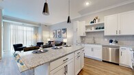 This thoughtfully designed 1-bedroom, 1-bathroom residence offers a comfortable and modern living experience across a single floor. The open-concept layout seamlessly connects the kitchen and living area, creating a bright and functional common space finished with warm wood-look flooring throughout.

The kitchen is a standout feature, showcasing crisp white shaker cabinets with matte black hardware, granite countertops, a subway tile backsplash, and stainless steel appliances including a refrigerator, range, and microwave. A central island with granite surface and pendant lighting above provides additional prep space and casual seating for multiple bar stools.

The living room is generously proportioned with crown molding, neutral gray walls, and a large sliding glass door fitted with vertical blinds that floods the space with natural light and provides outdoor access.

The bedroom is a serene retreat featuring plush carpet, large windows with outdoor views of greenery, and ample space for a full furniture arrangement. The bathroom is well-finished with a granite-topped white vanity, wood-look flooring, and a shower-tub combination. Throughout the residence, consistent design details — including granite surfaces, white cabinetry, and black hardware accents — lend a cohesive and modern aesthetic.