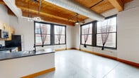 Located at 1110 Washington Ave in the heart of downtown St. Louis, this 2-bedroom, 2-bathroom loft showcases a striking industrial aesthetic with authentic architectural character. The open-concept living, dining, and kitchen area is anchored by soaring exposed wood-beam ceilings with visible ductwork and a polished concrete floor that flows throughout the entire space. Large, black-framed windows flood the interior with natural light and frame views of the surrounding urban skyline.

The kitchen is well-appointed with light maple cabinetry, dark countertops, stainless steel appliances including a refrigerator, range, microwave, and dishwasher, and a central island with a sink — ideal for both cooking and casual dining. A ceiling fan with track lighting adds to the functional yet stylish feel of the main living area.

Both bedrooms continue the loft's design language with concrete floors, exposed beam ceilings, and generous windows offering city views. The spacious primary bedroom includes an en-suite bathroom. Each bathroom features wood-toned vanities with dark countertops, white tile tub-shower combinations, and black hardware accents. Additional storage is provided by four closets throughout the unit. A narrow hallway with exposed ceiling joinery connects the private areas of the home.