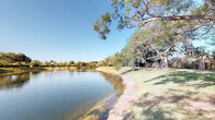 This single-storey property is distinguished primarily by its exceptional outdoor setting. The surrounding landscape features direct access to a calm, reflective waterway, with a gently sloping grassy bank leading to the water's edge. Mature trees — including broad canopy varieties — provide natural shade along the shoreline, contributing to a peaceful and private atmosphere. The water view stretches across to a tree-lined opposite bank, with glimpses of neighbouring properties visible in the distance, suggesting a well-established residential environment. The clear sky above and the stillness of the water create a serene, park-like ambiance that defines the character of this space. Unfortunately, interior images are insufficiently lit to confirm specific details regarding room layouts, finishes, or architectural features inside the property. What is evident from the exterior perspective is that the natural surroundings serve as a significant and defining asset, with the waterfront position and mature landscaping providing a tranquil backdrop that would be visible from various points within the home.