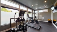 Located on SW Orbit Street in Beaverton, Oregon, this fitness center is a thoughtfully designed single-floor space built to support a comprehensive workout experience. The interior features a neutral color palette of warm taupe and dark gray tones, complemented by recessed lighting and durable rubber flooring throughout.

Large windows line the walls, allowing abundant natural light to fill the space while providing views of the surrounding neighborhood. Privacy film on the lower panes balances openness with discretion. The layout is open and well-organized, providing ample room to move between stations.

The cardio section is equipped with multiple True-brand machines, including treadmills, an elliptical, a recumbent bike, and a spin bike. A dedicated strength area features a multi-tier dumbbell rack with a full range of weights, medicine balls, a workout bench, and a mat for floor exercises. A beverage refrigerator provides added convenience for hydration during workouts.

A motivational "Fit, Fierce, Fabulous" wall graphic serves as a focal point, adding an energetic character to the space. A wall-mounted screen and posted fitness center rules round out the functional details. The overall ambiance is clean, modern, and purpose-driven, making it a practical and inviting space for daily fitness routines.