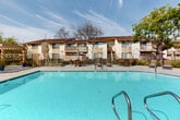 Located at 1059 Casitas Pass Rd in Carpinteria, California, this outdoor community area serves as a central gathering space defined by its generously sized swimming pool with crystal-clear blue water. The pool is accessible via stainless steel entry rails and is enclosed by a secure metal fence, ensuring both safety and privacy for residents. Surrounding the pool is a spacious concrete deck outfitted with lounge chairs, chaise lounges, and dining-style seating, providing ample room for relaxation and socializing under the Southern California sun. A substantial wood-beam pergola on one side of the pool deck offers welcome shade and a covered outdoor lounge area, complemented by decorative planters with vibrant tropical greenery. Adjacent to the pergola is a single-story clubhouse or amenity building, accessible via a ramp for ease of access. The landscape is enriched by several mature trees that frame the space and provide natural shade throughout the day. The surrounding multi-story residential buildings feature private balconies that overlook the pool area, creating a cohesive community environment. The overall ambiance is relaxed and resort-like, reflecting the mild, pleasant climate characteristic of Carpinteria's coastal setting.