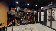 Located on E Roosevelt Rd in Chicago, this single-floor space is designed with a distinctly music-centric identity. The most striking feature is a floor-to-ceiling installation covering an entire wall with an eclectic collection of vintage and modern speakers, audio equipment, suitcases, and sound gear arranged in a dense, curated collage. Recessed spotlights set against a dark ceiling illuminate this display, creating a dramatic focal point that immediately sets the tone for the space.

The interior design blends dark, moody aesthetics with bold graphic elements. One area features cork wall and floor tiles arranged in a large geometric chevron pattern, contrasting warm natural tones with dark panels. Industrial-style bar stools and wooden seating add functional character. Another section showcases bold skull-patterned wallpaper paired with a mounted flat-screen television and soft bean bag seating, contributing a lounge-like atmosphere. Decorative metallic wall accents and directional lighting complete this area's edgy, artistic vibe.

Underfoot, patterned area rugs soften the space alongside smooth flooring. A glazed interior door featuring a headphones logo hints at dedicated studio or listening room functionality. Throughout, the design balances raw, vintage character with intentional artistic detailing, creating an immersive environment suited for music-focused activities.