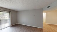 This single-story space offers a practical and cohesive layout spread across several well-defined areas. The living room is a standout feature, showcasing wood-look luxury vinyl flooring in a neutral gray tone and a textured ceiling that adds dimension overhead. A full-width sliding glass door fitted with vertical blinds floods the room with natural light while providing outdoor access. An adjacent area with matching flooring connects seamlessly to the rest of the unit, demonstrating a consistent design throughout the common spaces.

The kitchen is notably updated, featuring dark wood-toned cabinetry with modern hardware, crisp white quartz-style countertops, and a classic white subway tile backsplash. A black electric range with an overhead microwave and ventilation hood anchors the cooking area, combining functionality with a contemporary aesthetic.

The bedrooms are carpeted in neutral tones, offering a softer underfoot feel, and are finished with the same light gray walls found throughout. One bedroom includes a window with blinds that allows for natural light and outdoor views. The bathroom includes a full tub and shower combination with chrome fixtures and a curved shower rod. Throughout the space, white baseboards and neutral wall tones create a clean, move-in ready environment.