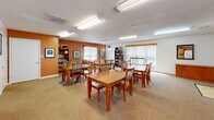 Located at 7910 Sterling Cir in Toano, Virginia, this single-floor community space is thoughtfully designed to serve as a shared gathering and dining area. The layout comprises two primary areas: a generously sized dining room and a dedicated kitchen, offering a practical and welcoming environment for group use.

The dining room is the heart of the space, featuring wall-to-wall carpet flooring and multiple wooden dining tables with matching ladder-back chairs, comfortably accommodating several groups simultaneously. Flush fluorescent lighting illuminates the room evenly, while large windows fitted with roller shades allow natural light to filter in. Warm terracotta-orange accent walls add a distinctive, inviting character throughout. A bookshelf and a cushioned sofa tucked into one corner provide a relaxed lounge area, complemented by framed artwork on the walls.

The kitchen is compact yet efficiently arranged, featuring warm oak cabinetry both above and below a continuous white countertop. A double-basin sink sits centrally along the main wall, and a full-size white refrigerator anchors one end of the layout. Light vinyl tile flooring and the same terracotta accent wall tie the kitchen's aesthetic seamlessly to the rest of the space. A window with horizontal blinds brings in additional natural light. The space also includes restroom access, making it well-suited for community events and gatherings.