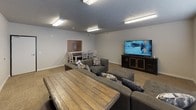 This community room is a versatile, single-floor shared space designed to foster social interaction and relaxation among its users. The room is finished with neutral gray walls and textured ceilings, creating a clean and understated aesthetic throughout. Durable carpet tile flooring in a warm beige tone covers the entire space, providing comfort underfoot.

The layout is thoughtfully organized into distinct zones. A generously sized lounge area anchors the room, furnished with a large gray sectional sofa and matching ottoman, accented with decorative throw pillows. A rustic wood coffee table sits at the center, adding a warm contrast to the neutral surroundings. Flush-mounted fluorescent light fixtures provide bright, even illumination throughout.

A flat-screen television mounted on a dark wood media console serves as the entertainment focal point, making the lounge ideal for group viewing. Adjacent to the lounge, a dining area features a dark wood table surrounded by white painted chairs, accommodating group meals or activities. A pass-through window connects this area to an adjoining space, enhancing the room's functionality.

A wide window with white horizontal blinds allows natural light to filter in, while a wall-mounted air conditioning unit ensures year-round comfort. A standard entry door provides convenient access, completing this practical and inviting shared community space.