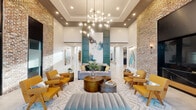 This thoughtfully designed single-floor amenity space combines upscale aesthetics with functional communal living. The grand lobby lounge commands immediate attention with soaring double-height ceilings, exposed brick accent walls, and a dramatic multi-globe chandelier. Mustard-yellow armchairs, a plush gray sectional, and layered area rugs create an inviting seating arrangement, while large-scale abstract artwork and teal architectural panels add bold visual interest. A built-in brick-framed media wall with an oversized flat-screen serves as a secondary lounge focal point. The communal kitchen and dining area features a generous white quartz island beneath statement brass pendant lights, stainless steel appliances, and a cozy banquette lounge with upholstered bench seating, marble-top tables, and curated wall art — ideal for casual gatherings. The expansive fitness center is exceptionally well-appointed, offering a full array of cardio machines, a dedicated free weights zone with floor-to-ceiling mirrors, and a separate functional training studio with herringbone wood-panel walls, hardwood floors, and suspension training equipment. Exposed brick, track lighting with dark-painted ceiling coffers, and large windows throughout the fitness areas bring in natural light and maintain design cohesion across the entire space.
