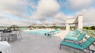Located along Big Bend Road in Twin Oaks, Missouri, this rooftop pool and amenity deck offers a well-appointed outdoor retreat designed for both relaxation and social gathering. The centerpiece of the space is a generously sized swimming pool with crystal-clear water, built-in entry steps, and multiple access points via stainless steel railings. The surrounding deck is finished in large-format light gray porcelain tile, providing a clean, modern aesthetic underfoot.

Rows of teal-cushioned lounge chairs line the perimeter of the deck, offering ample space for sunbathing with open sky above. A glass railing system runs along the outer edges of the rooftop, ensuring unobstructed panoramic views of the Twin Oaks landscape while maintaining a secure boundary. Several pergola structures with string lighting provide shaded dining and seating areas, furnished with white tables and chairs suited for casual outdoor meals or conversation.

A distinctive hanging egg chair adds a stylish lounge nook near the pool's edge. The space is accessed through a glass-panel door set within a modern tile-clad exterior wall. The overall design is contemporary and cohesive, combining functional amenities with elevated outdoor living in an open-air rooftop setting.