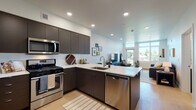 This single-floor residence showcases a thoughtfully designed open-concept layout that seamlessly connects the kitchen, dining, and living areas. The kitchen is a standout feature, equipped with sleek dark-finish cabinetry, white quartz countertops, stainless steel appliances including a gas range, built-in microwave, dishwasher, and refrigerator, and a peninsula island that provides additional workspace and casual seating. Light wood flooring runs throughout, complementing the clean white walls and recessed lighting overhead.

The living area is bathed in natural light from large floor-to-ceiling windows that frame outdoor neighborhood views, creating a bright and welcoming atmosphere. A dedicated bedroom continues the light-filled aesthetic, featuring a generously sized window with scenic views and ample space for furnishings. A closet with sliding doors and built-in shelving provides practical storage.

The bathroom is finished in a minimalist style, featuring dark-toned cabinetry, a white quartz vanity, an illuminated mirror, and light-toned tile flooring. An entry area with an accessible closet and electrical panel rounds out the functional layout of this well-appointed modern residence.